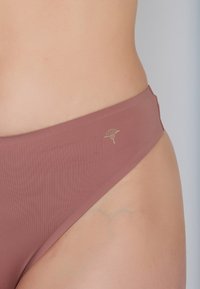 Mauve bikini bottom with smooth fabric, high-cut design, and subtle ribbed texture. Features a gold logo accent on the side.
