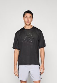 Μαύρο αθλητικό μπλουζάκι με κοντά μανίκια από δίχτυ, με εμφανές ασημένιο λογότυπο Adidas στο μπροστινό μέρος. Φοριέται με ανοιχτό γκρι σορτς.