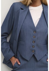 Dopasowany niebieski garnitur z usztywnioną marynarką z klapą z wycięciem oraz dopasowaną kamizelką z pięcioma brązowymi guzikami i bocznymi kieszeniami.