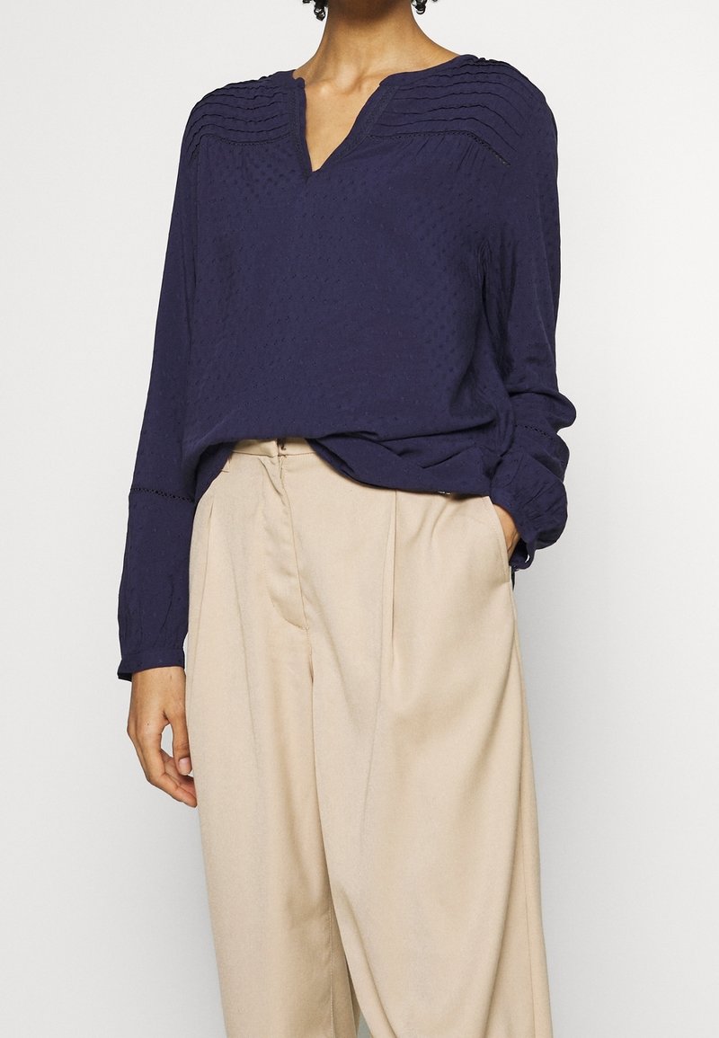 Blouse bleu marine texturée avec col en V, manches longues et plis horizontaux. Associée à un pantalon beige ample.