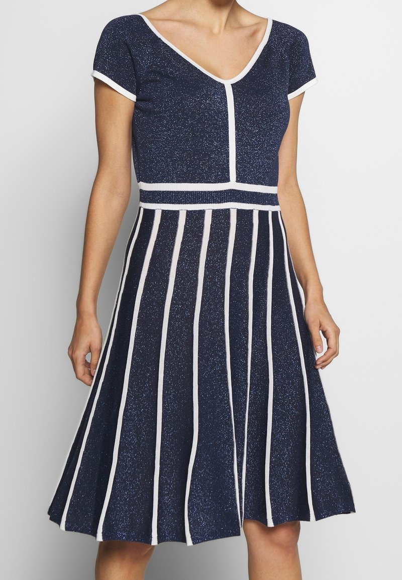 Mörkblå stickad klänning med korta ärmar, V-ringning, figursydd överdel och utsvängd kjol med vita vertikala ränder och glittrig textur.