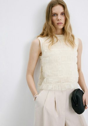 Mujer con cabello largo y ondulado, vestida con una blusa sin mangas de encaje color crema y pantalones beige claros, sosteniendo un bolso de mano negro tejido sobre un fondo blanco.