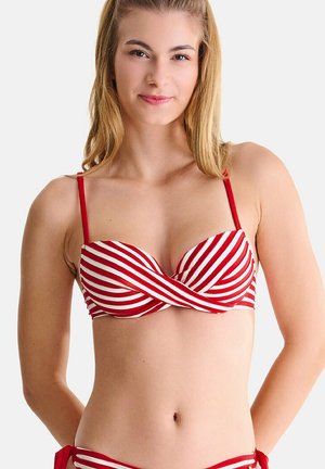 Jeune femme portant un haut de bikini rouge et blanc à rayures avec des fines bretelles rouges sur un fond clair uni.