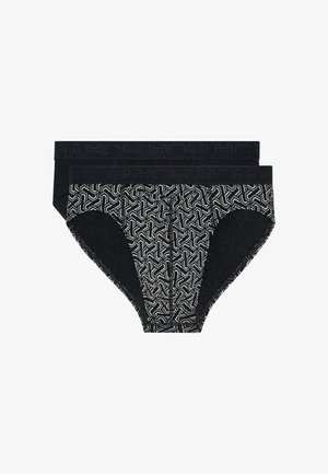 Gespierde man staat op blote voeten, draagt zwarte slip met patroon, kijkt naar zijn linkerzijde tegen een effen lichte achtergrond.