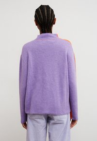 Personne aux cheveux tressés portant un pull lilas avec des rayures orange sur les manches et un pantalon en velours côtelé violet clair, tournée dos à l'appareil photo.