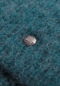 Tkanina wełniana w odcieniu teal o miękkiej fakturze z srebrnym guzikiem, na którym umieszczony jest tekst "TY" w nowoczesnej czcionce, położonym centralnie.