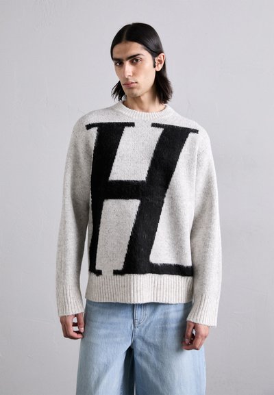 Suéter de punto gris claro con una gran "H" negra en la parte frontal. Puños y dobladillo acanalados, textura suave y corte relajado. Combinado con jeans azules.