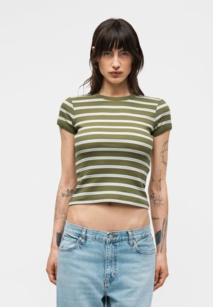 Kobieta o ciemnych włosach nosi zielono-białą, paskowaną krótką koszulkę i jasnoniebieskie dżinsy, prezentując tatuaże na obu ramionach na jednolitym tle.