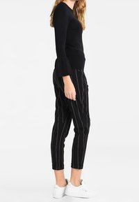Czarny top z długim rękawem i rozkloszowanymi mankietami połączony z czarnymi spodniami w paski i białymi trampkami. Spodnie mają pionowe, białe prążki.