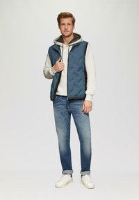 Veste rembourrée bleu marine avec une fermeture éclair, portée sur un sweat à capuche gris. Jean bleu clair et baskets blanches complètent le look. Texture lisse, design décontracté.