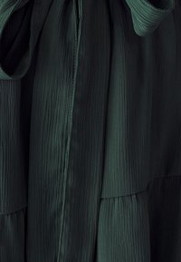 Langes, dunkelgrünes Kleid aus strukturiertem, plissiertem Stoff. Es hat ein Bindeband in der Taille, eine glatte Oberfläche und vertikale Nähte.