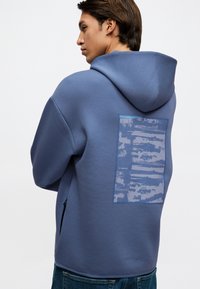 Νεαρός άνδρας με μπλε φούτερ με κουκούλα, με ψηφιακό ορθογώνιο γραφικό στην πλάτη, στέκεται σε προφίλ μπροστά σε άσπρο φόντο.