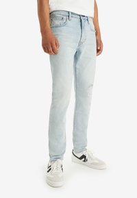 Mies, joka pukeutuu vaaleansinsisiin kapeisiin farkkuihin, joissa on pieni kulutusmerkintä oikeassa reidessä, ja mustilla yksityiskohdilla varustettuihin valkoisiin tennareihin, seisoo valkoista taustaa vasten.