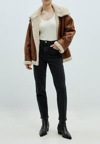 Braune Jacke mit Schaffellfutter und beigem Kragen, kombiniert mit hochgeschnittenen schwarzen Jeans und schwarzen Stiefeletten mit Absatz.