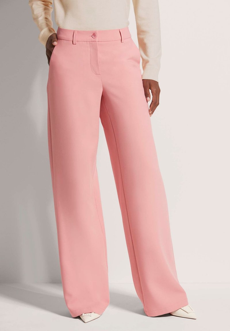 Roze wijde broek met een gladde textuur, voorzien van een knoopsluiting en zijzakken. Gecombineerd met puntige witte schoenen.