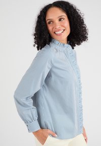 Lichtblauwe en wit gestreepte blouse met een hoge kraag, knoopsluiting en een gerimpelde accent langs de voorkant. Lange mouwen met manchetten.