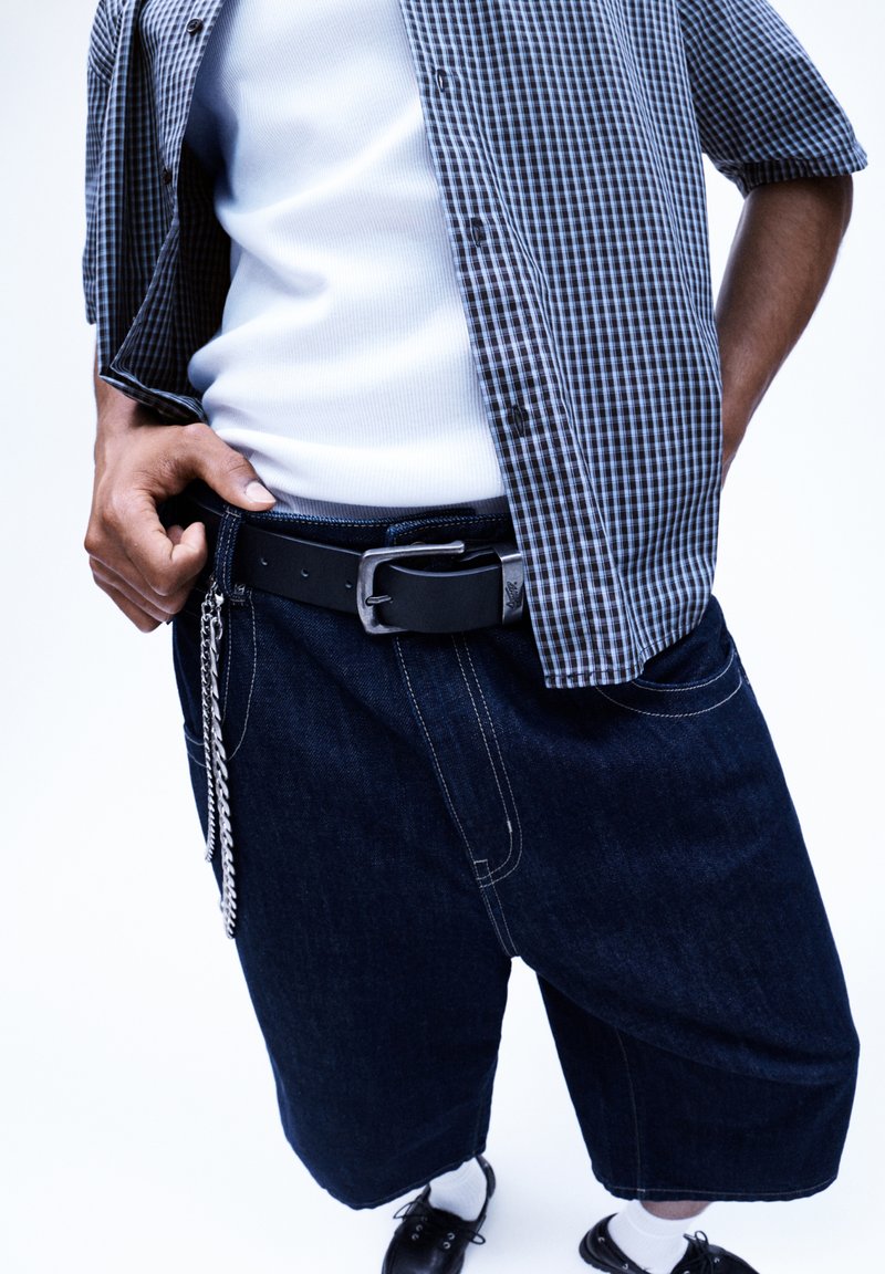 Donkerdenim shorts met een losse pasvorm, gecombineerd met een zwarte riem met een metalen gesp en een zilveren kettingdetail. Geruit overhemd met knopen.