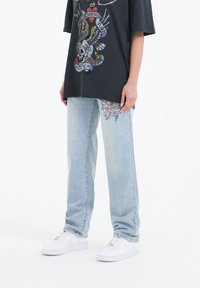 T-shirt nera con grafica di teschio colorato e ali, abbinata a jeans denim blu chiaro con design floreale e sneakers bianche.
