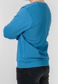 Blå bomulls sweatshirt med långa ärmar, rund hals och ribbad nederkant. Texturerad tyg synlig, bärs med svarta jeans.