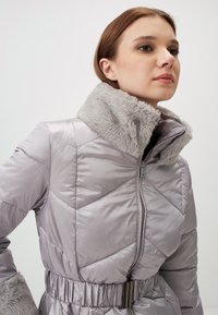Giacca imbottita grigio chiaro con colletto in pelliccia, caratterizzata da un motivo a rombi diagonali e una vita elastica. Chiusura con zipper frontale e tasche visibili.