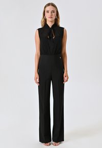 Combinaison noire avec un design sans manches, un col à nouer et une taille haute. Le tissu est lisse, structuré et présente un pantalon à jambes larges.