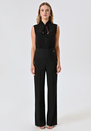 Schwarzer Overall mit ärmellos Design, Bindekragen und hoher Taille. Der Stoff ist glatt, strukturiert und verfügt über weit geschnittene Hosenbeine.