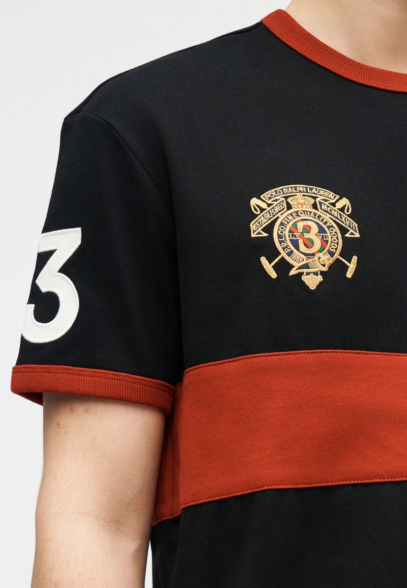 Chemise à manches courtes noire et rouille avec écusson brodé sur la poitrine et numéro blanc 3 sur la manche droite.