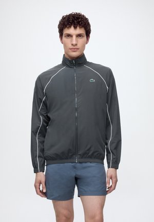 Homme aux cheveux bouclés portant une veste zippée gris foncé avec des liserés blancs et un short bleu, debout devant un fond blanc uni.