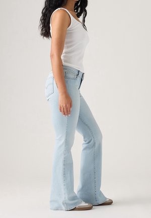 Frau trägt hellblaue ausgestellte Jeans, weißes Tanktop und weiße Sneakers, steht seitlich vor einem schlichten Hintergrund.