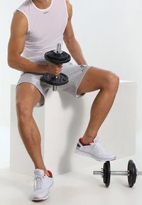 Top grigia senza maniche, pantaloni corti grigi e sneakers bianche con accenti rossi. Tiene un manubrio nero. Sfondo geometrico semplice.