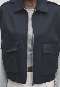 Granatowa kurtka bomberka z frontowym zamkiem błyskawicznym, dwiema kieszeniami naszytymi, elastycznym pasem i gładką fakturą, noszona na szarym topie.