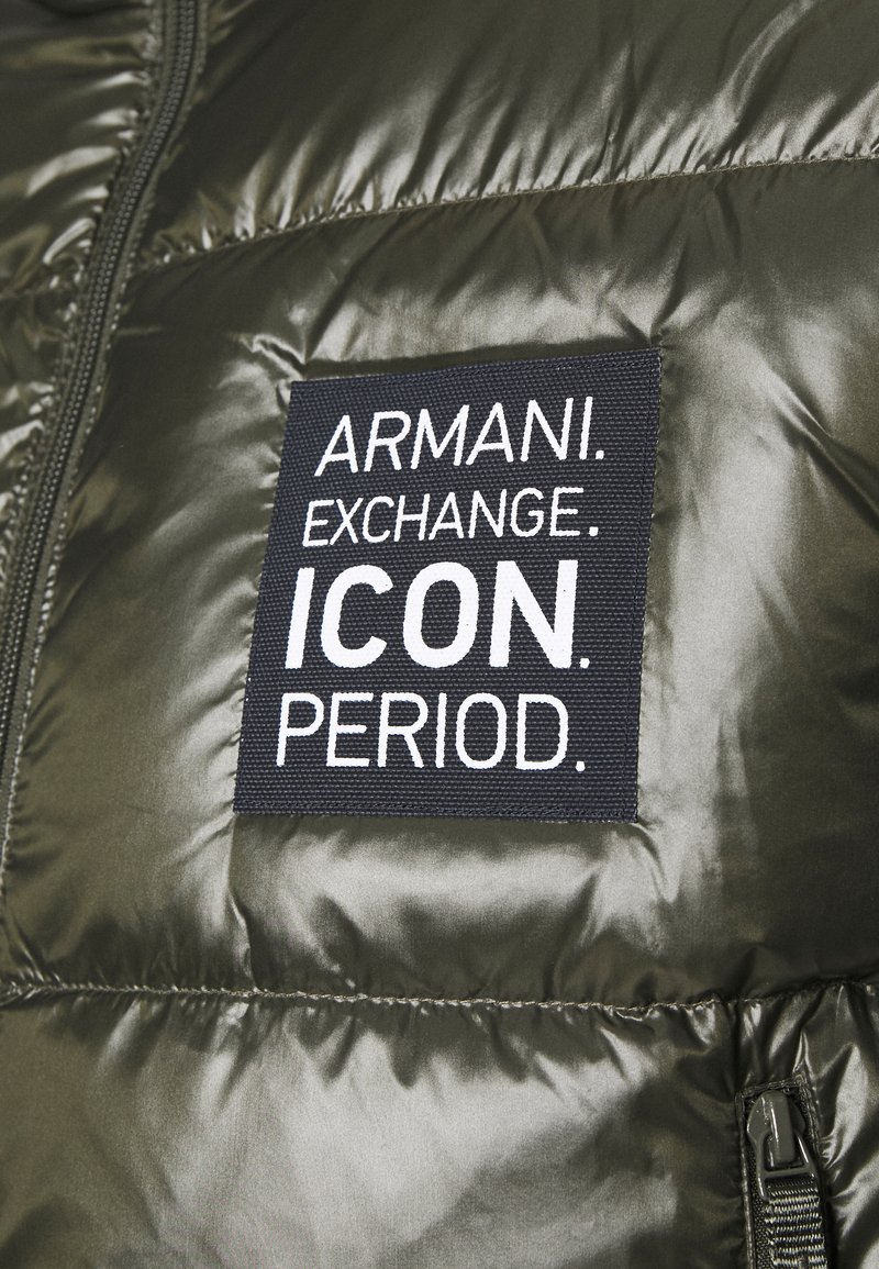 Giacca puffer verde oliva con una superficie liscia, caratterizzata da un'etichetta nera ben visibile che riporta la scritta "ARMANI. EXCHANGE. ICON. PERIOD." in testo bianco.