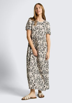 Vrouw staand, gekleed in een beige enkellange jurk met zwarte bloemenprint en korte pofmouwen, gecombineerd met beige instapsandalen.