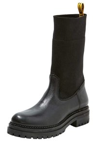 Svart ankelboot med en slät lädertå och häl, ribbad stickad ovandel och en kraftig gummisula med mönster. Gul dragflikdetalj.