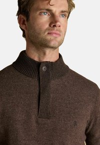 Brauner Strickpullover mit hohem geripptem Kragen und Knopfverschluss. Verfügt über eine feine Textur und ein kleines Logo auf der Brust.