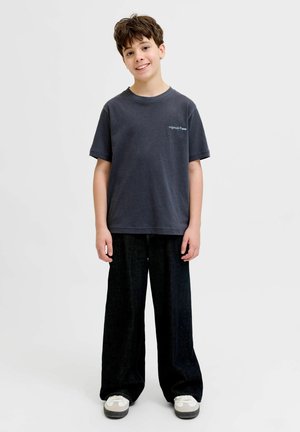 Smaidošs jauns zēns stāv, tērpies tumšā pārlieku lielā T-kreklā, plašās melnās biksēs un baltos sporta apavos, uz gaiša vienveidīga fona.