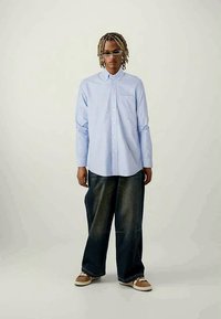Lichtblauwe overhemdd met lange mouwen en een voorzak, gecombineerd met losse donkere denim broeken en sneakers. Het model staat voor een neutrale achtergrond.