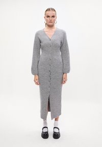 Abito grigio lavorato a maglia con scollo a V, maniche lunghe a palloncino, chiusura frontale con bottoni e spacco all'orlo. Accentuato da scarpe nere e calzini bianchi.