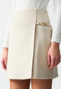 Beige suède mini rok met een wikkelontwerp en een gouden sluiting aan de zijkant, met een gladde textuur en strakke lijnen.