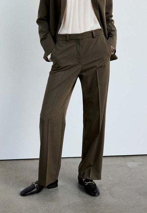 Personne portant un pantalon marron sur mesure, des mocassins noirs avec une boucle dorée, une chemise blanche et un blazer marron, debout les mains dans les poches.