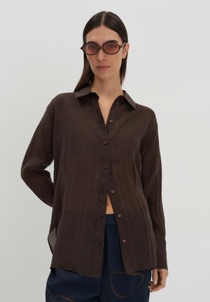 Chemise marron à boutons avec un tissu doux et léger, présentant un col, des manches longues et des fentes sur les côtés pour faciliter les mouvements.