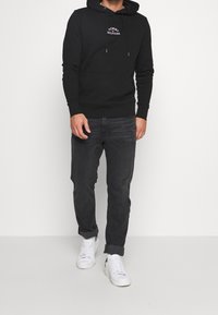 Black hooded sweatshirt with white logo print, paired with dark jeans and white sneakers. The fabric appears soft, with ribbed cuffs and hem.