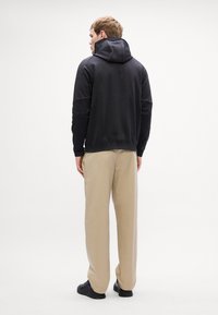 Črna kapuca s črnim elastikastim robom, v kombinaciji s svetlo bež hlačami. Gladka tkanina z minimalnimi šivi, obuta v črne čevlje.