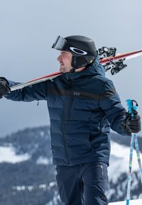 Skijacke in Dunkelblau mit dem oberen Abschnitt in Schwarz, kombiniert mit Skihosen. Trägt einen schwarzen Helm und hält Skier über der Schulter.