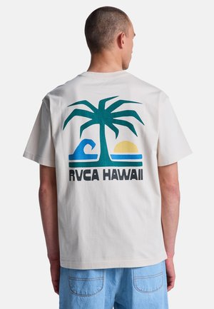 Man draagt wit T-shirt met een teal palmboom, blauwe golf, gele zon en de tekst "RVCA HAWAII" op de achterkant, gecombineerd met lichtblauwe spijkershorts.