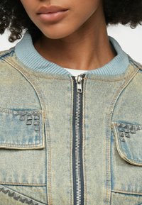 Modra denim jakna z svetlo modrim rebrastim ovratnikom, zadrgo in kovinskimi žebljički na žepih. Mehka tekstura in sproščen oblikovanje.