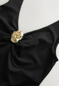 Tissu noir torsadé au centre avec un détail de fermoir métallique doré brillant et irrégulier sur un vêtement sans manches.