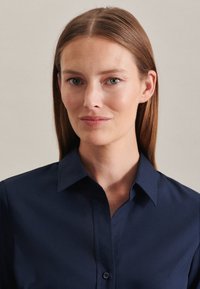 Camicia a colletto navy realizzata in tessuto liscio, con un design a bottoni e una silhouette aderente. Leggero lucido evidente sulla superficie.