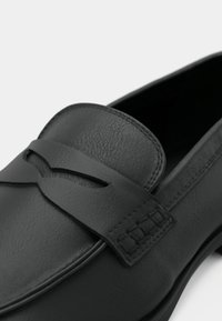 Mocassin en cuir noir avec une bande décorative et un design ajouré sur le dessus. Présente des détails de couture le long des bords et un bout arrondi.