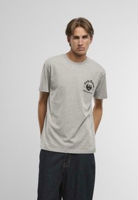 Tricou gri cu mânecă scurtă și guler rotund, prezentând o imprimeu grafic negru pe partea stângă a pieptului, confecționat dintr-un material moale din bumbac.